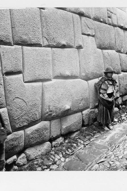 Black and white photograph from around 1907 of a woman in a wide hat and shawl standing barefoot against a wall of enormous tightly fitted polygonal Inca stone blocks on a cobbled Cuzco street.