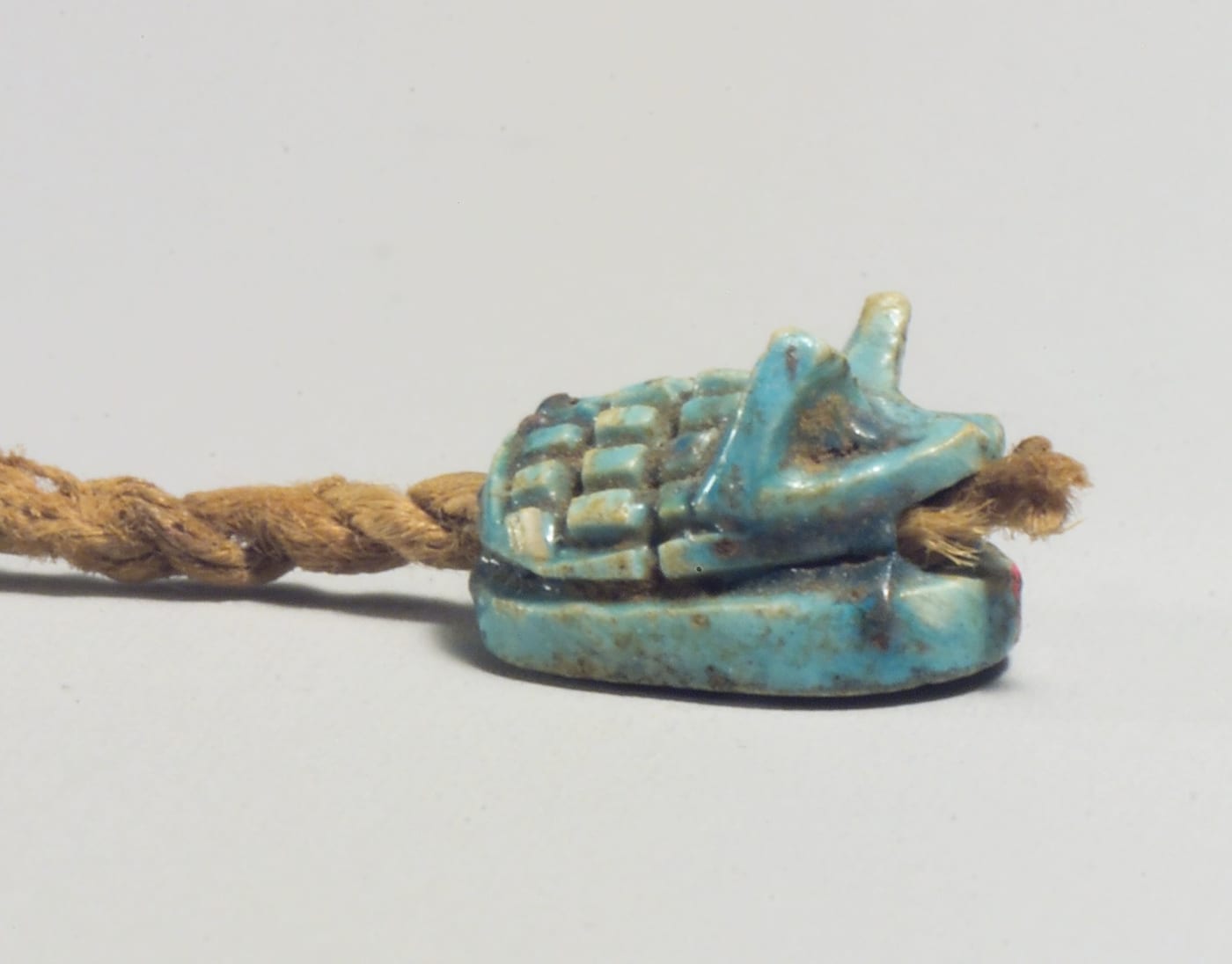 A small turquoise glazed steatite amulet in the form of a hedgehog, its spines carved as a raised grid across the back, resting on an oval base and threaded onto a frayed linen cord.