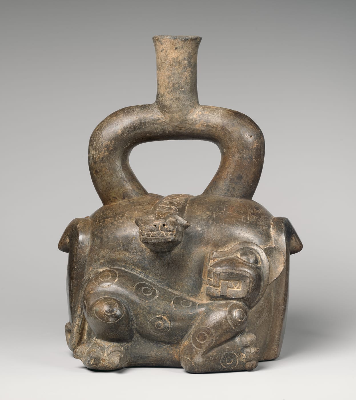 A dark burnished ceramic stirrup-spout bottle modelled in low relief with a fanged feline whose body is covered in concentric ring markings, alongside a stepped fret design.