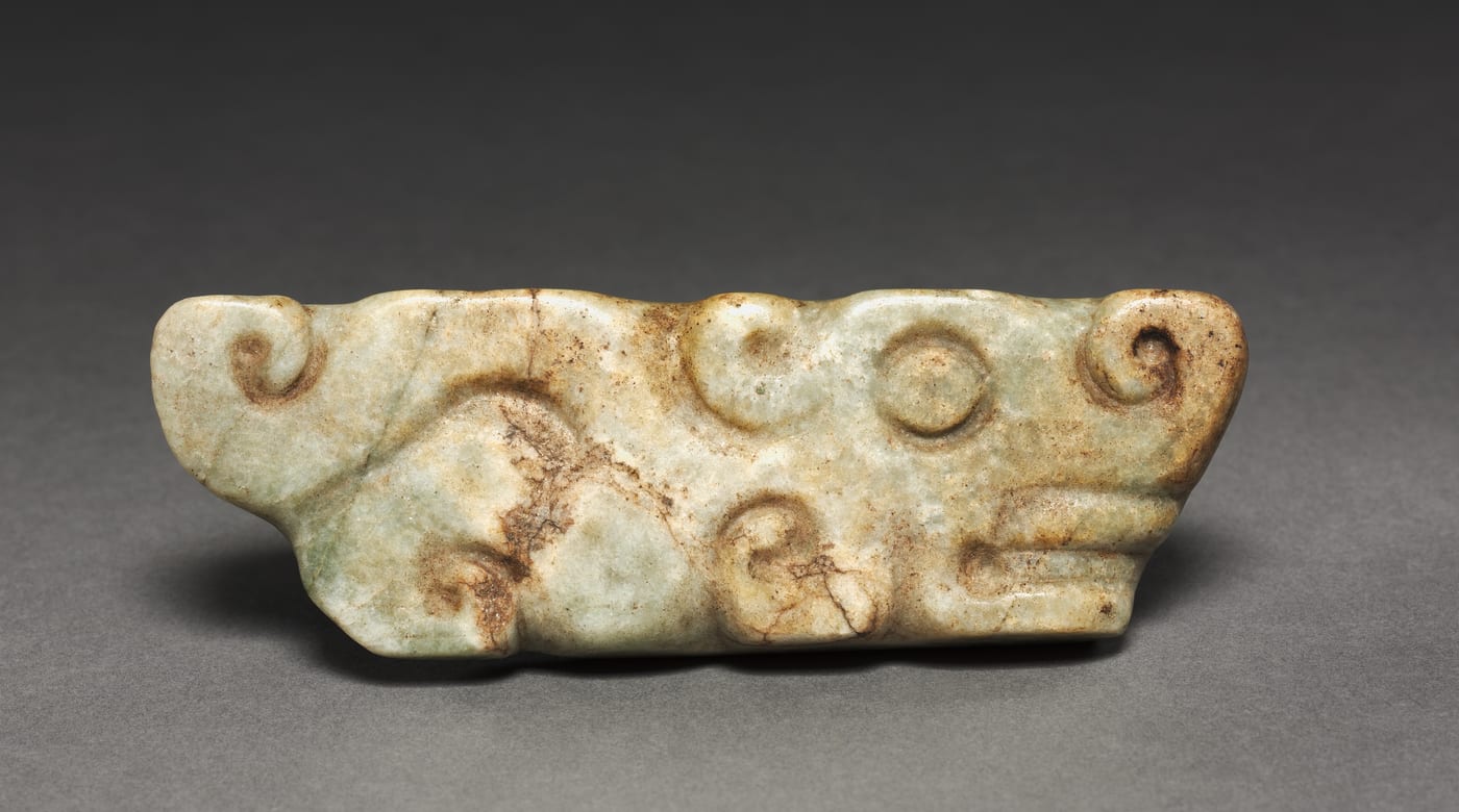 A pale green jade plaque carved in low relief with scroll forms, a ringed eye and a curled snout with a projecting jaw, identified as a jaguar pendant.