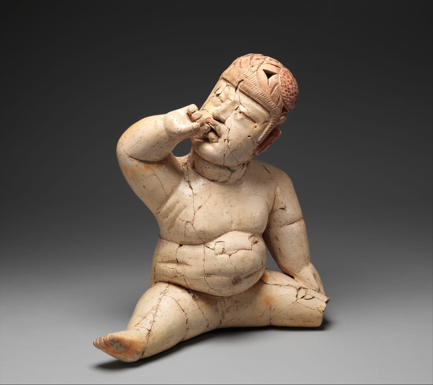 Olmec hollow ceramic figure of a seated infant with a heavy rounded body, one hand raised to its face, wearing a close-fitting helmet, the mouth turned sharply down at the corners.