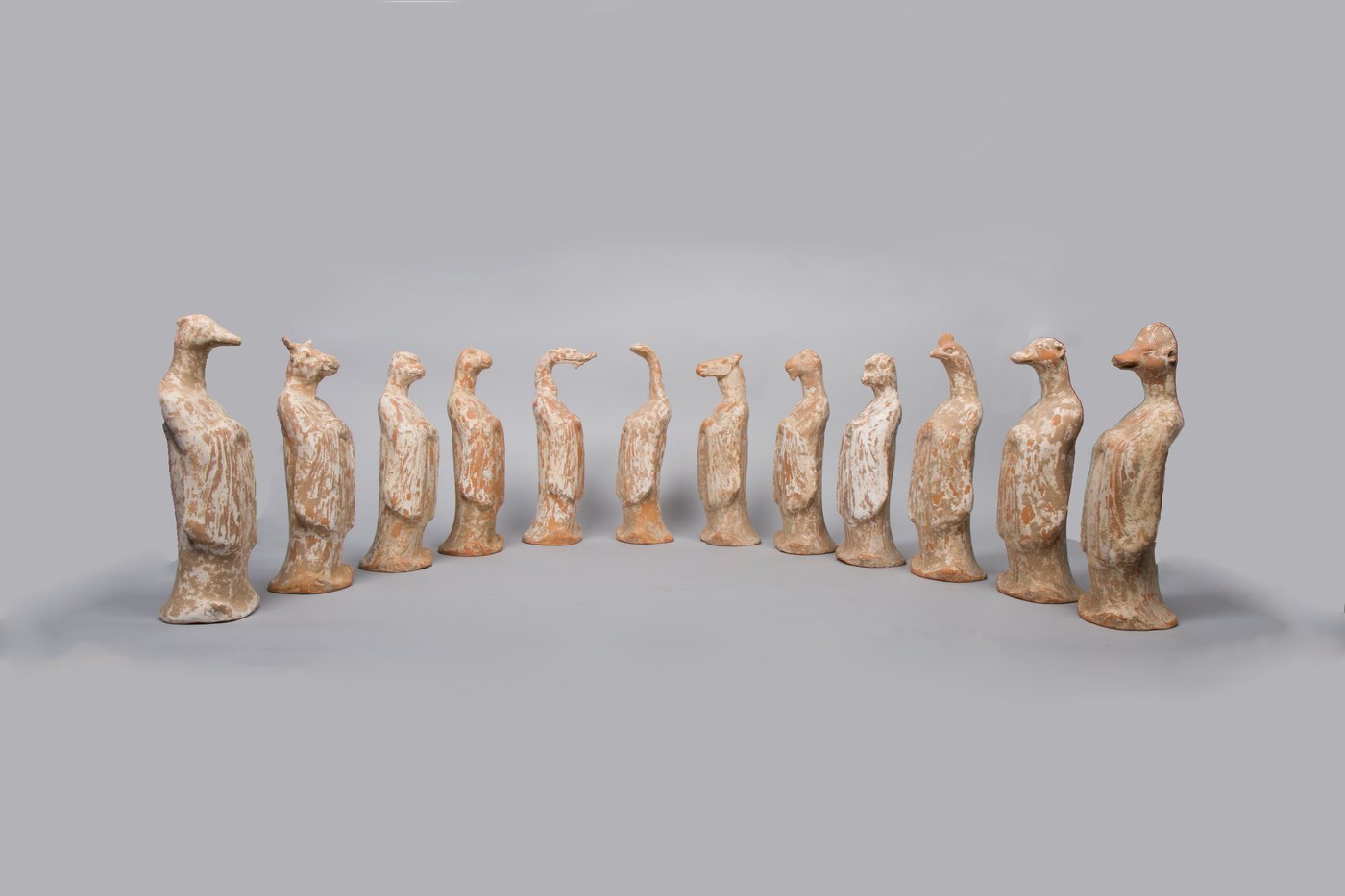 A row of twelve small Tang dynasty earthenware figures with human bodies in long robes and different animal heads, arranged in an arc against a plain grey ground.
