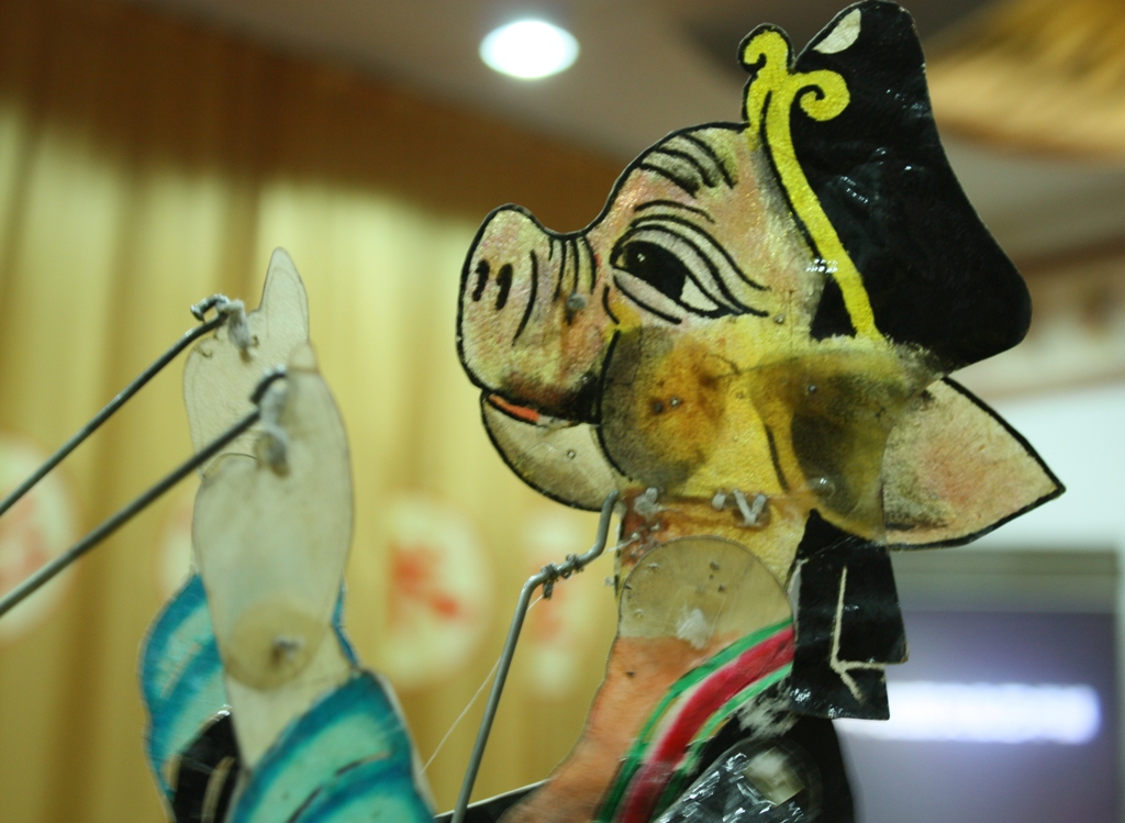 Chinese shadow puppet of Zhu Bajie, a translucent painted leather figure with a pig's head, long snout and large ears, wearing a hat and robes, held on control rods.