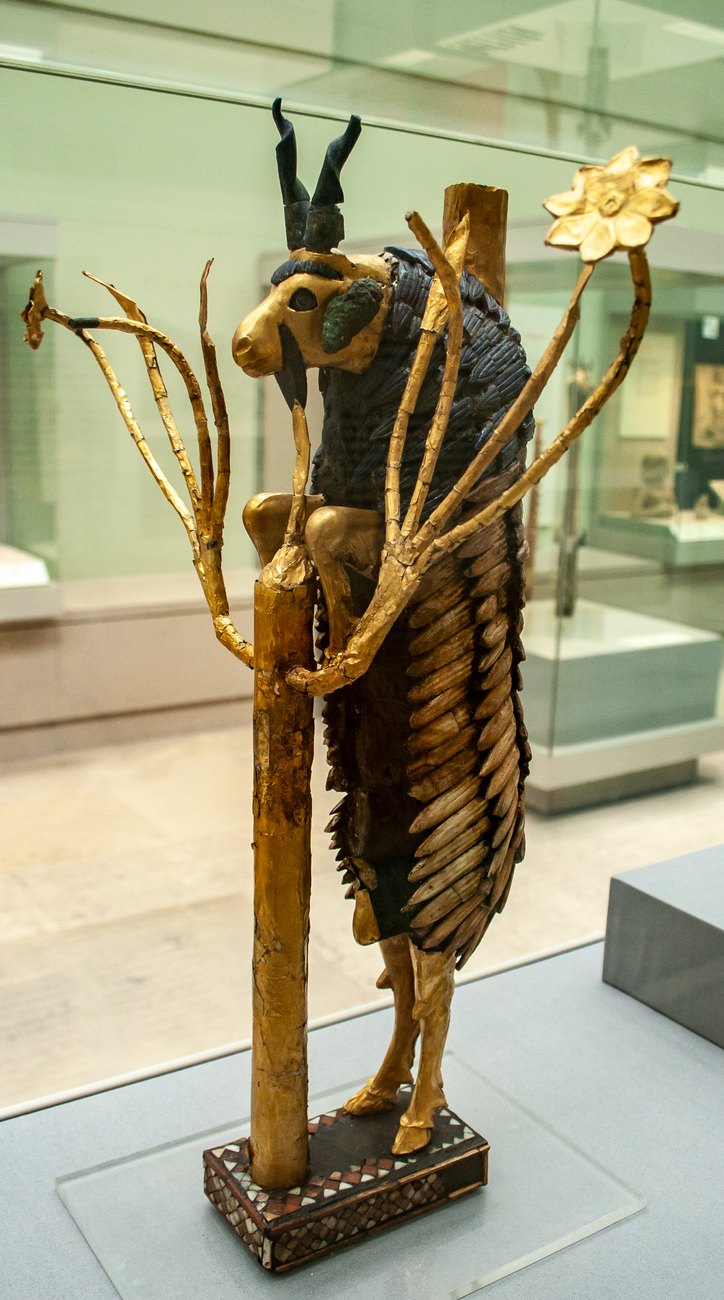 The Ram in a Thicket statuette from Ur displayed in a museum case: a gold-headed caprid with blue lapis horns and shoulder fleece rearing against a golden plant with a flower at the top.