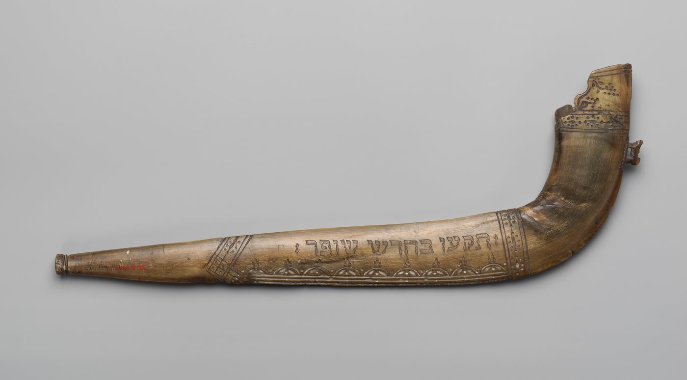 An engraved shofar, a long upturned animal horn with a Hebrew inscription and decorative bands incised along its length, photographed against a plain grey ground.