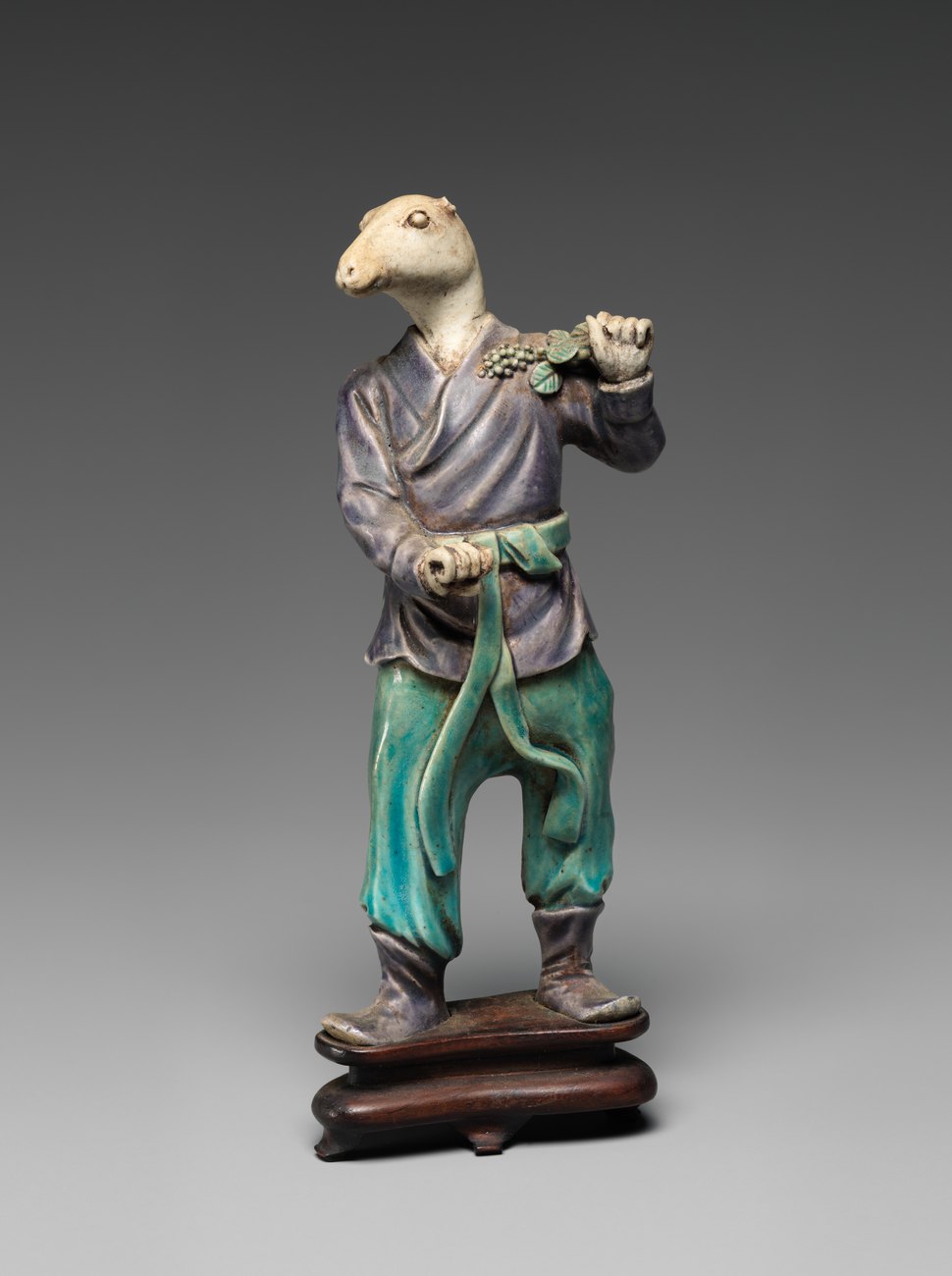 Chinese porcelain figure of a standing man in purple and green robes with the head of a rat, mounted on a carved wooden stand.