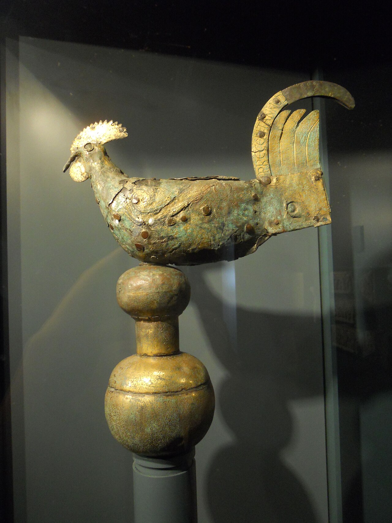 Ninth-century copper weathercock with a raised comb and a broad upswept tail, pitted with age, mounted on a bronze sphere in a museum display case.